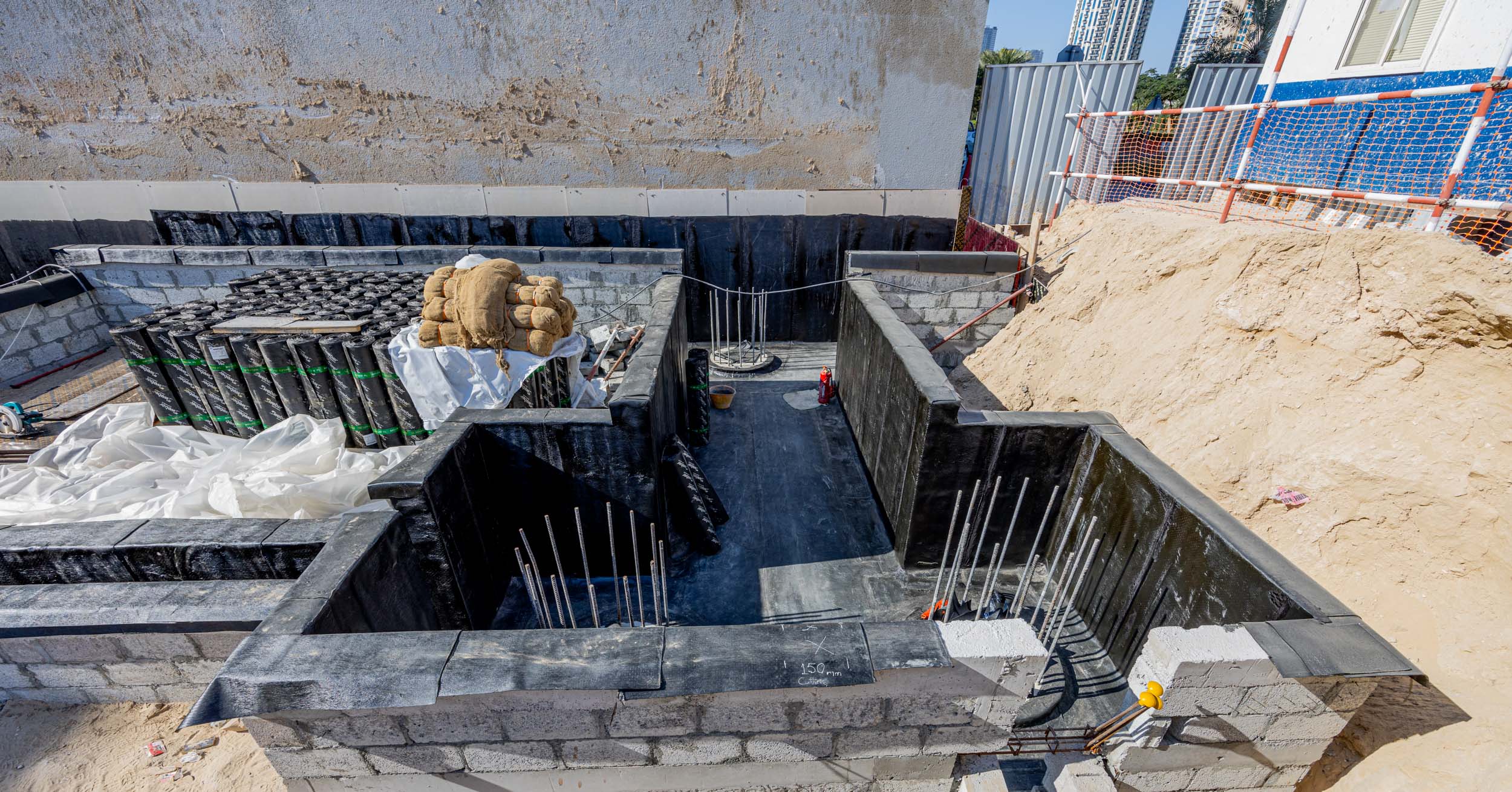 Pearl House By Imtiaz Construction site featuring a foundation pit with concrete blocks, waterproofing material, and exposed rebar.